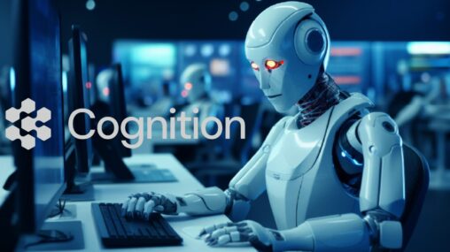 A plastic humanoid robot awash in a glow of blue screen sits at a desk and stares somewhat dully at a computer screen on the left It has one hand resting on a keyboard.